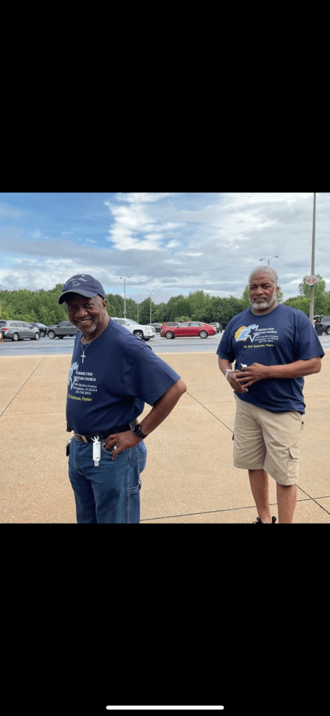 Two Men Wearing Blue Shirts Stand Together in a Parking Lot