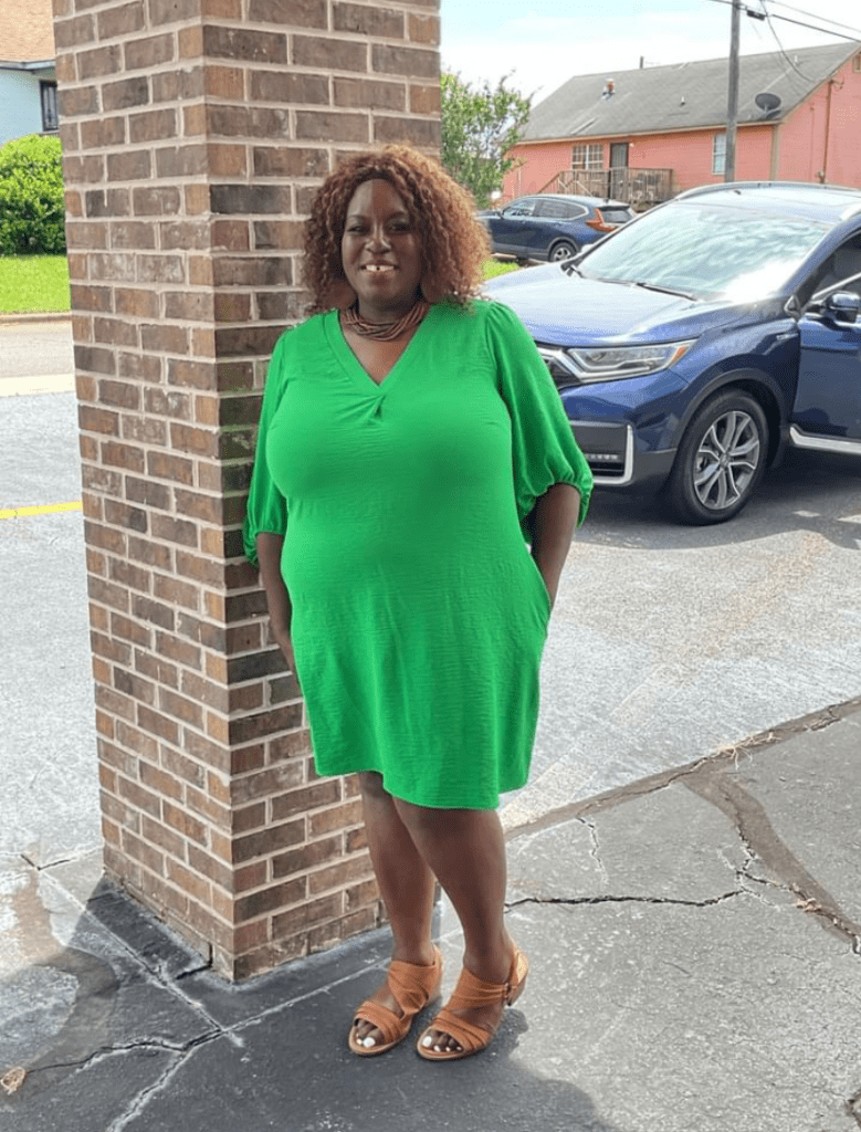 A Woman in a Green Dress Stands Gracefully Beside a Textured Brick Wall
